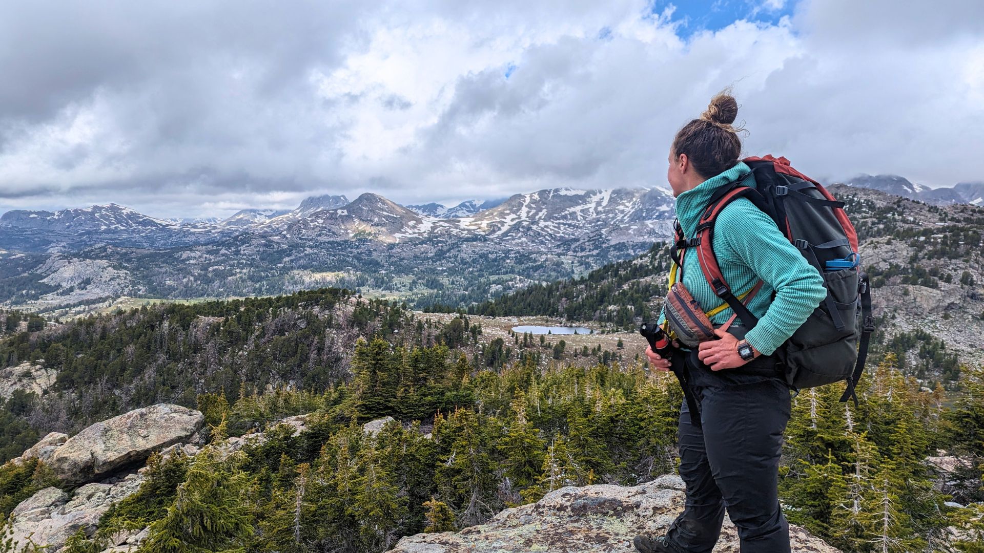 A woman with a bearspray and a backpack on stands looking out at the wind river mountains